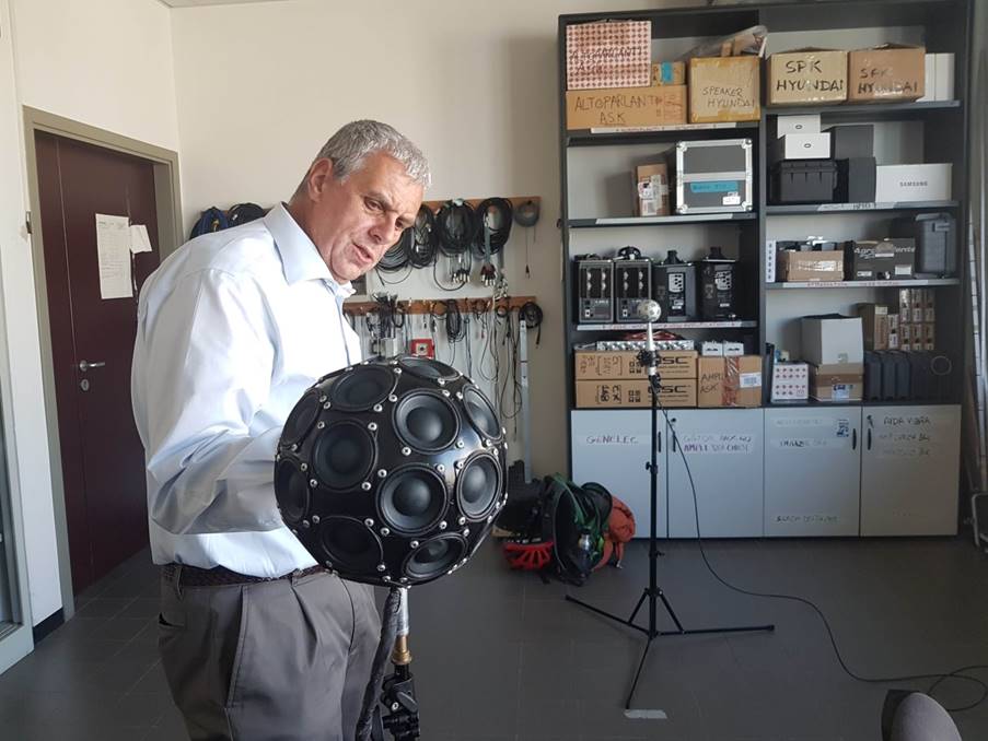 A gray-haired man stands between a black sphere, which is a loudspeaker array, and a silver sphere, which is a microphone array.