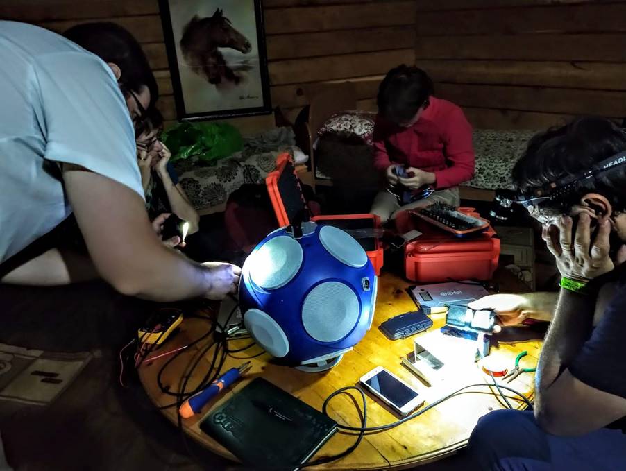 Three people surround the dodecahedron in a dark room, peering at the speaker array with torches.